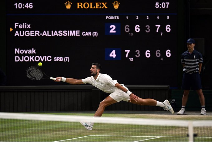Novak Djokovic, seeded (7) from Serbia, stretches for a forehand volley during his match against Canada's Felix Auger-Aliassime (3), with the scoreboard showing Djokovic leading 4 sets to 2 in a five-set battle. The Rolex-branded scoreboard reflects the promotional focus on "Djoker," highlighting Djokovic's competitive performance throughout the match.