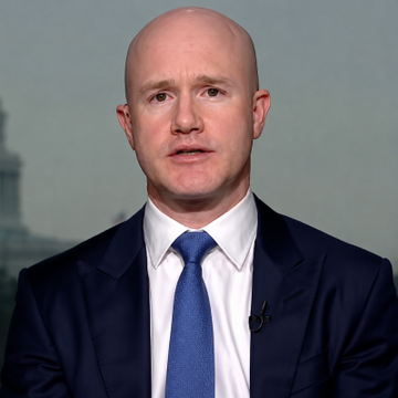 A bald man in a dark navy suit and blue tie speaks during what appears to be a televised interview, with a lapel microphone visible on his jacket.