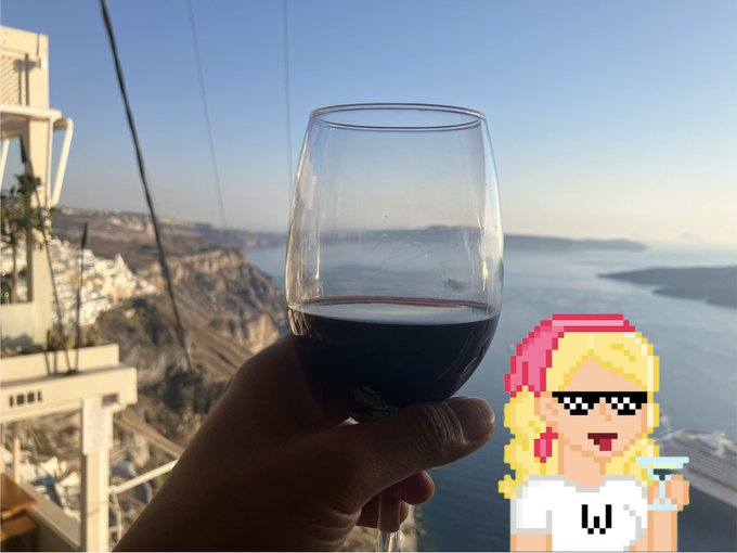A hand holds a glass of red wine against the breathtaking backdrop of Santorini's caldera, with its iconic white-washed buildings and blue Aegean Sea. A pixel art character wearing sunglasses and holding a cocktail adds a fun, playful touch to the scenic toast, perfectly capturing the celebratory spirit of "Giamas" (Greek for "Cheers").