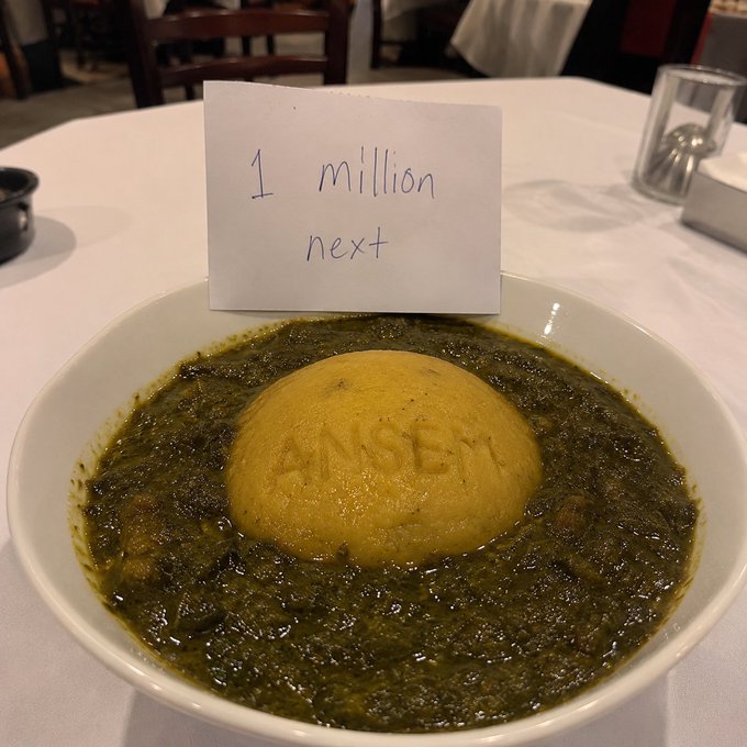 A white bowl holds a round mound of yellow Eba (with "ANSEM" embossed on it) surrounded by green stew, with a handwritten sign reading "1 million next" stuck into the dish. The image playfully stages the Eba as a branded "$ANSEM" dish, tying into the post's theme of celebrating the meme coin's milestone and aiming for 1 million holders.