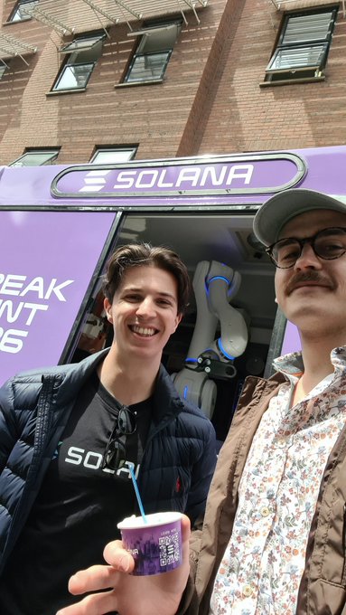 Two men smile for a selfie in front of a purple Solana-branded bus, with one wearing a Solana shirt and holding a small purple cup with a straw.