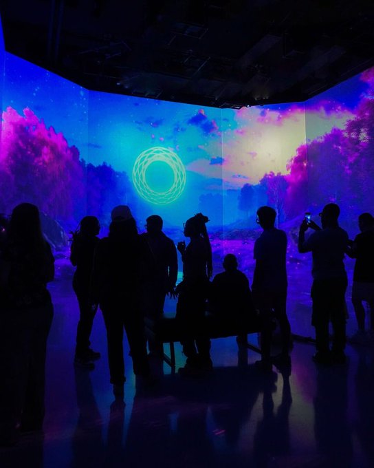 Silhouettes of visitors gaze at and photograph a large circular cyan projection surrounded by purple and blue cosmic clouds in an immersive digital art exhibition. The display aligns with "SUBMERGE" at Artechouse DC, showcasing works like "Satori" by J.Pierce, "LATTICE" by MhxAlt, and "The River" rendered in 18K resolution.