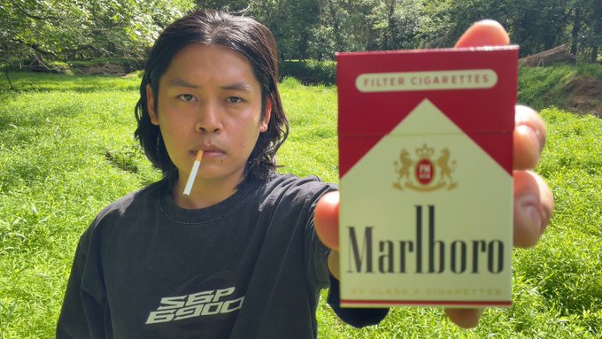 The image features a young man holding up a Marlboro cigarette box, with his hand positioned over it as if he's about to light it or has just finished smoking one. The background of the photo shows a grassy field, which adds a natural element to the scene. The caption at the bottom of the image reads "Smoking a cigarette, believing in spx690? and singing an original at 4pm Watch: youtu.be/1Kd6M6z15nM?".
