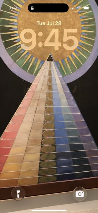 An iPhone lockscreen displaying "Tue Jul 28" and "9:45" over artwork featuring a triangular pyramid composed of horizontal bands in various colors, set against a circular sunburst background. The image visually matches the post's sentiment of cherished long-term use, suggesting the user has kept this striking, mystical artwork as their lockscreen for six years.