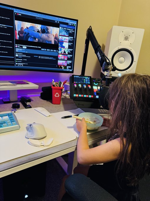 A person sits at a desk with a bowl of food, watching YouTube on a monitor, surrounded by streaming equipment including a microphone on a boom arm, an audio mixer, a studio monitor speaker, and various desk accessories.