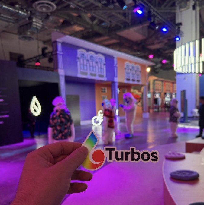 A hand holds a small card at an event with an overlaid 'Turbos' logo, while costumed characters including Stitch appear in the background under colorful lighting.