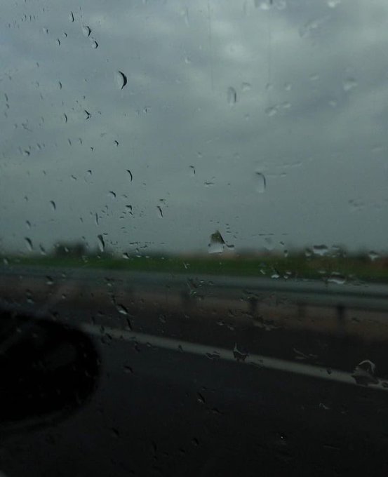 A moody view through a rain-covered windshield shows a gray, overcast sky and a highway with blurred vehicles in the distance.