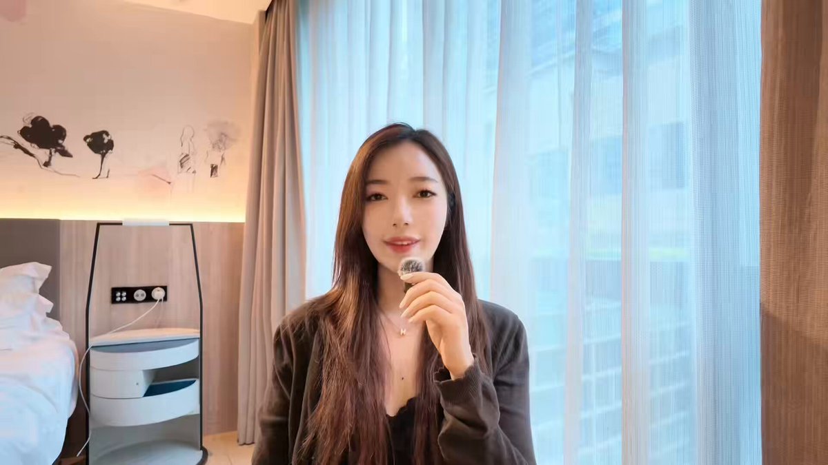 The image features a woman with long brown hair holding a microphone and smiling at the camera while standing in front of a window. The background includes curtains and a bed, suggesting that she might be in her bedroom or a hotel room. The caption provided is related to the stock market crash, which has been widely discussed on social media platforms like Twitter. It seems that the woman's smile could be interpreted as an attempt to spread positivity amidst the negative news about the stock market crash.