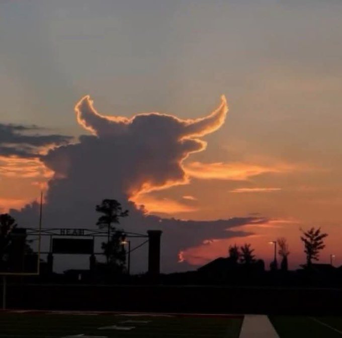 A silhouette of a football field at sunset features a dramatic cloud formation that resembles a horned skull or demon face in the orange sky above.