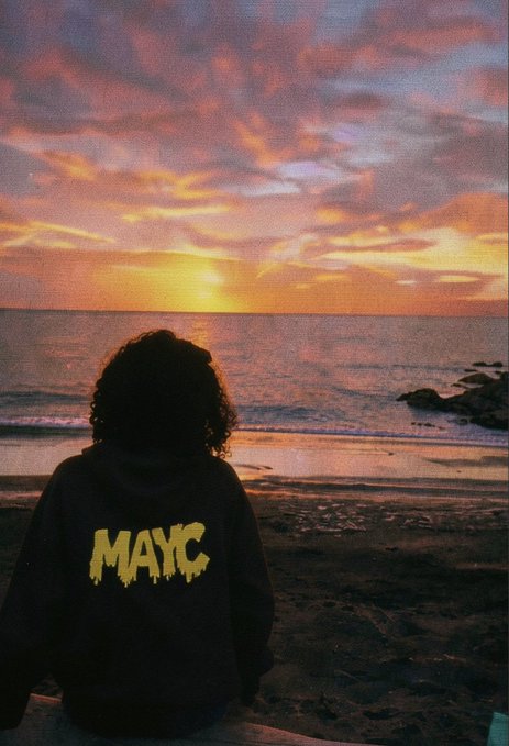 A person with curly hair wearing a black hoodie with yellow 'MAYC' text sits on a beach facing a colorful sunset over the ocean.