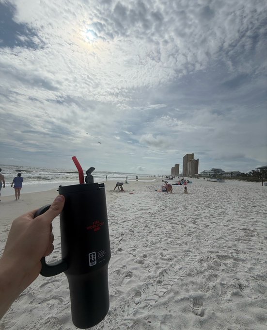 A hand holds up a black tumbler with a red straw against a beach scene, featuring white sand, scattered beachgoers, and high-rise buildings in the distance under a partly cloudy sky.
