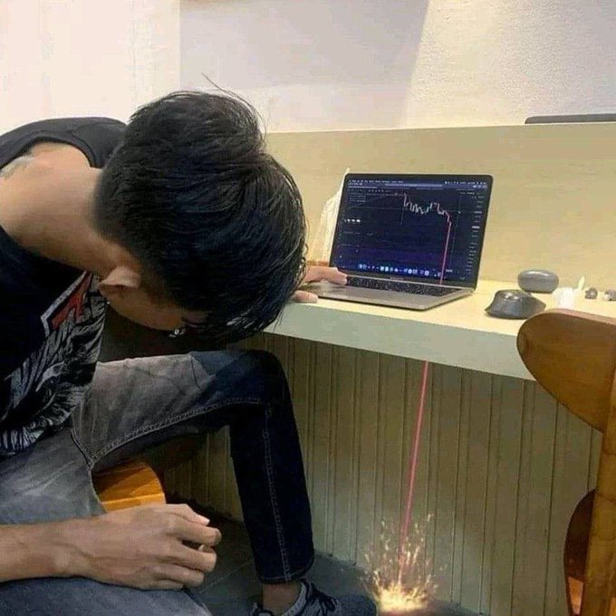 A person slumped over a desk in front of a laptop displaying a declining trading chart, with a red line shown dramatically plunging off the desk.