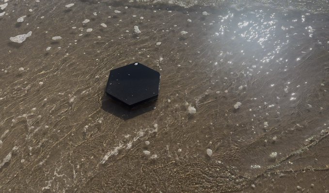 A black hexagonal device resting in shallow, foamy seawater on a sandy beach.