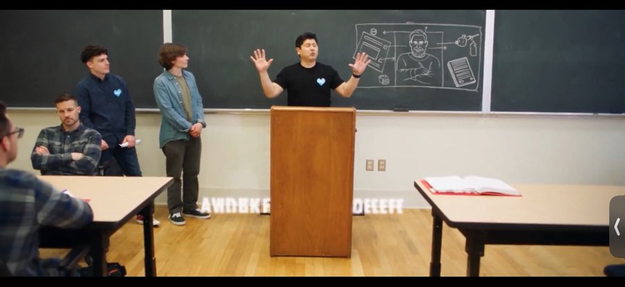 A photo of a presentation in a classroom where a man at a wooden podium gestures enthusiastically, with three other men standing behind him near a chalkboard featuring drawings.