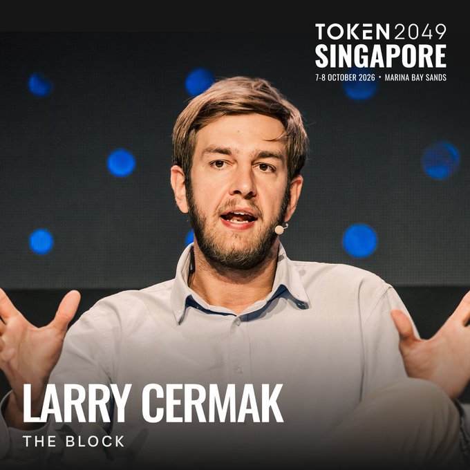 A bearded man in a light button-down shirt speaks on stage, gesturing with both hands, with blue bokeh lights in the dark background. The promotional graphic announces "TOKEN 2049 SINGAPORE, 7-8 OCTOBER 2026 • MARINA BAY SANDS" and identifies the speaker as "LARRY CERMAK, THE BLOCK," tying the image to his return appearance at the crypto conference.