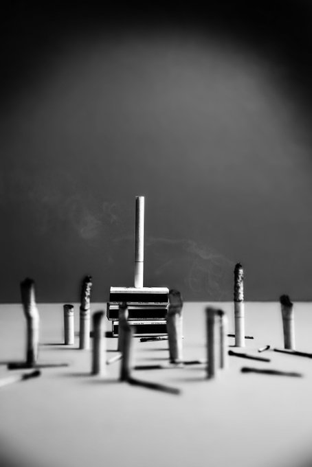 A black and white photograph shows a close-up of several razors standing upright on a surface with wisps of smoke in the background.