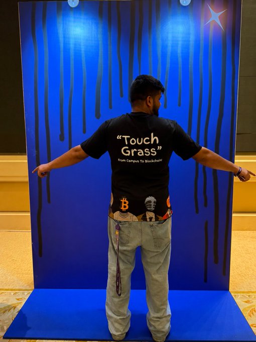 A man stands with arms outstretched in front of a blue backdrop with black drip designs, wearing a black 'Touch Grass - From Campus To Blockchain' t-shirt and jeans showing a Bitcoin and Satoshi Nakamoto belt.