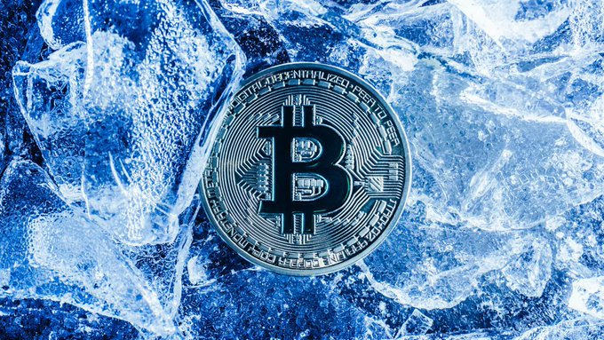A silver Bitcoin (₿) coin sits atop a bed of clear ice crystals against a blue-tinted background.