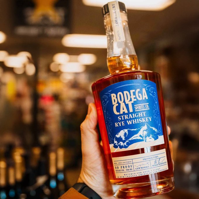 A hand holds up a bottle of Bodega Cat Spirits Co. Straight Rye Whiskey, featuring a blue label with a cat illustration, in front of a blurred store background.