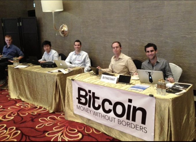 Five men sitting at a table with laptops behind a banner reading 'Bitcoin MONEY WITHOUT BORDERS', with a nameplate showing 'BITNSTANT'.