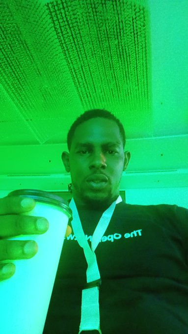 A man takes a selfie holding a white cup beneath a textured ceiling bathed in green light, wearing a lanyard and a shirt that reads "www.[blockchain].ent," apparently responding to a question about "Grammers" on the @ton_blockchain platform.