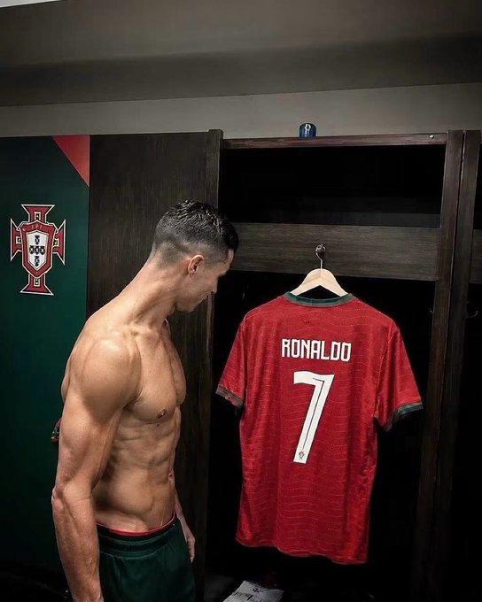 Cristiano Ronaldo stands shirtless in a locker room, displaying his physique next to a hanging red Portugal jersey with "RONALDO 7" on the back, with the Portuguese federation emblem visible on the wall.