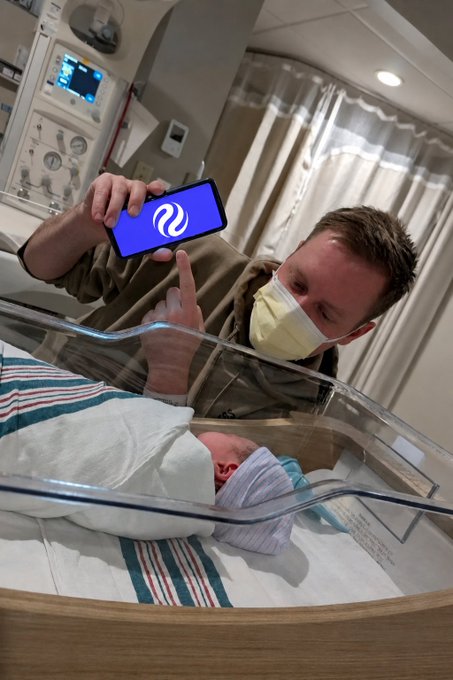 A masked father in a hospital leans over a newborn swaddled in a striped blanket inside a clear bassinet, proudly displaying his smartphone screen showing the Injective (INJ) crypto logo to the camera. The image visually represents the post's message of holding INJ as a long-term gift for his child, symbolizing early investment in the newborn's future.