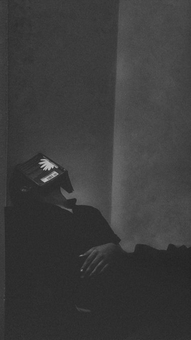 A dark, moody black-and-white image shows a person reclining in a shadowy room, wearing a hat with a glowing white hand or shell design illuminated on it.