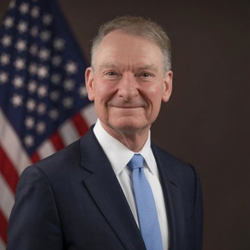 A portrait of an older man in a dark suit and blue tie smiling in front of an American flag.