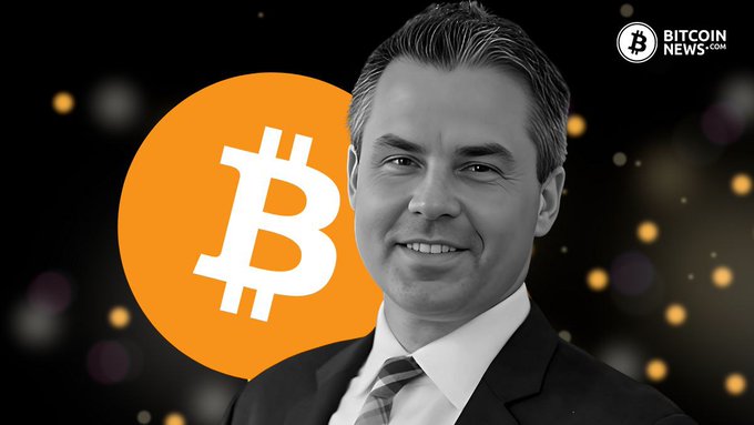 In the black and white photo, a man wearing a suit with his arms folded stands against a dark background. The man has short hair and is smiling at the camera. In front of him is an orange logo that features a stylized version of the letter "B". Below the man's head, there's a text stating "BALCHUNAS: $IBIT saw its second-largest daily outflow yesterday with - IBIT is still up over $2B YTD (top 2%) and has taken in roughly $64B in lifetime inflows. Yesterday's outflow was less than". The man seems to be a businessman, possibly involved in the cryptocurrency world or the Bitcoin news industry.