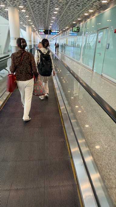 Two women walk on a moving walkway in a modern airport or transit terminal, carrying bags and luggage.