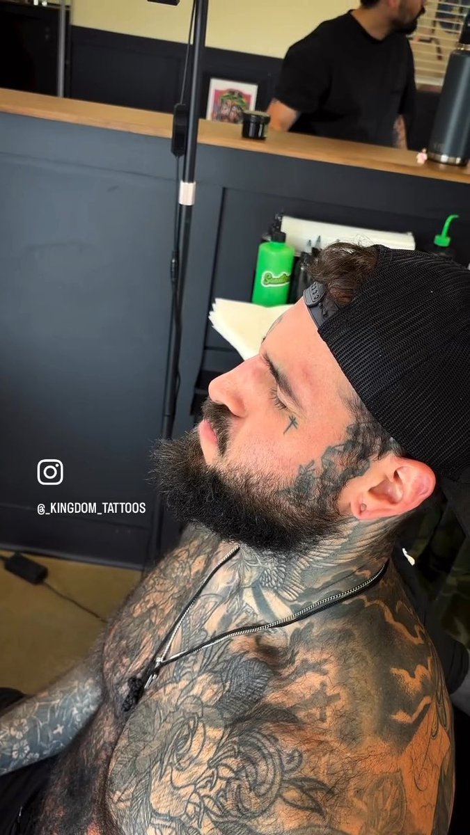 A bearded man with a black headband sits in a tattoo studio, his chest and neck covered in detailed tattoos including a cross under his eye, with the Instagram handle '@_KINGDOM_TATTOOS' displayed.