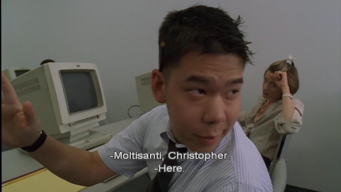 A young man in a striped shirt and tie raises his hand in a classroom with old CRT computers, captioned '-Moltisanti, Christopher. -Here.'
