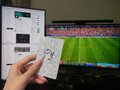 The image depicts a person holding up an electronic device that appears to be a remote control or gaming controller. Behind the device, there are two TVs showing a soccer match between different teams. The sports event on the televisions captures the attention of the viewer as they seem to observe the players closely.