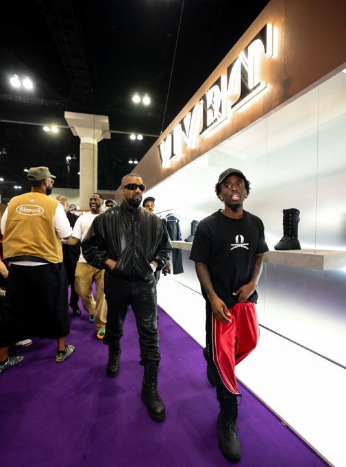 Kanye West and another person walk through a fashion or retail event space with a purple carpet, passing an illuminated 'LVRN' sign.