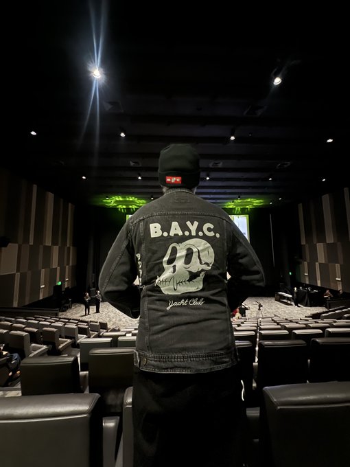 A person standing in a theater aisle wearing a black beanie with a YouTube logo and a denim jacket labeled 'B.A.Y.C.' with a skull graphic and 'Yacht Club' text.