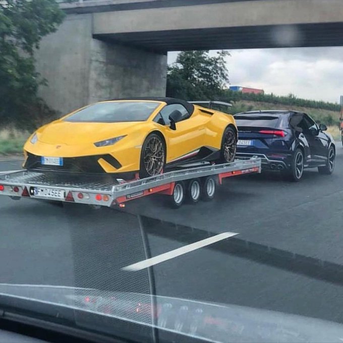 A yellow Lamborghini sports car is being transported on a flatbed tow truck on a highway, being pulled by a black SUV passing under a bridge.