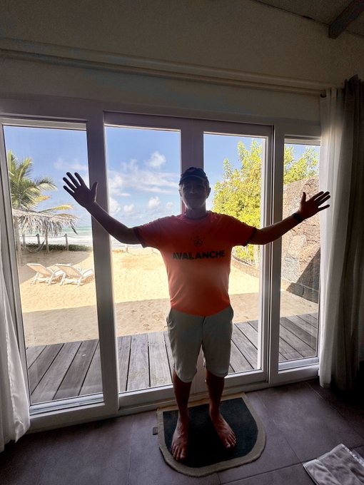 The image features a man standing in front of a window that opens onto a beach view. He has his arms outstretched as if he's embracing the beautiful scenery outside. The photo also includes text, which reads "Gbusiness Avax! Might be traveling with the family for the next week, but I am working hard everyday leading up to the launch of @YourGrails." This suggests that the man might be taking a break from work or other responsibilities and enjoying the view while doing so.