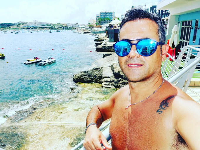 A shirtless man wearing blue reflective aviator sunglasses taking a selfie by a pool overlooking a coastal city with turquoise water and boats below.