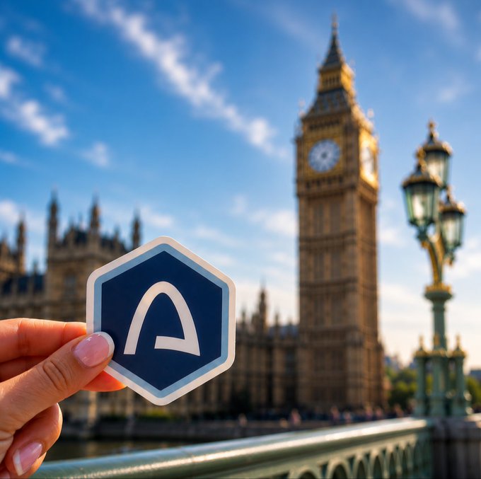 A hand holding a hexagonal token with a stylized 'A' logo in front of London's Big Ben, suggesting a UK-based crypto or blockchain project.