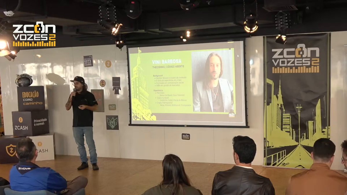 The image shows a man standing at a podium giving a presentation with a screen behind him displaying his face, possibly from a video conference or live streaming platform. The room has white walls and a wooden floor, creating an organized and professional setting for the event. There are several people in the audience, some sitting on chairs while others stand around the room. A banner is visible in the background that reads "Zcon Vozes 2".