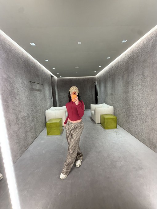 A woman takes a mirror selfie in a sleek, modern lounge wearing a pink cropped top, patterned Gucci-style pants, and a matching cap, captioned "Wifi money" — the upscale, minimalist interior with designer fashion conveying a flex of luxury and success tied to online income.