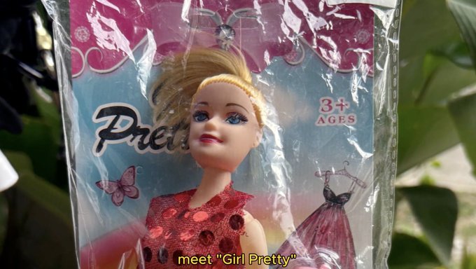 A close-up of a small doll in a red dress still in plastic packaging labeled "Pretty," with on-screen text reading 'meet "Girl Pretty"'.