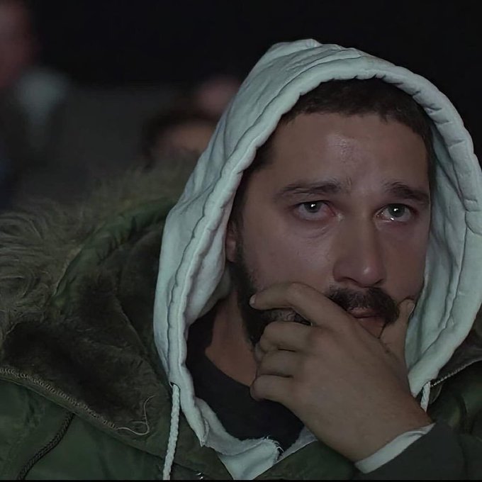 A close-up of actor Riz Ahmed with a contemplative expression, wearing a hooded jacket, appearing emotional with slightly watery eyes.