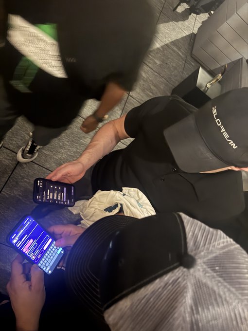 Two people wearing caps sit together looking at their smartphones, which display what appears to be trading charts with blue interfaces.