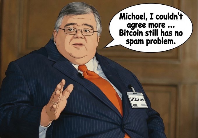 A cartoon-style illustration of a man in a pinstripe suit with an orange tie, wearing a "UTXO set" badge, says "Michael, I couldn't agree more... Bitcoin still has no spam problem." The satire mocks the idea of UTXO set growth as "spam" on the Bitcoin network, echoing GrassFedBitcoin's commentary on Bitcoin's block space usage debates.