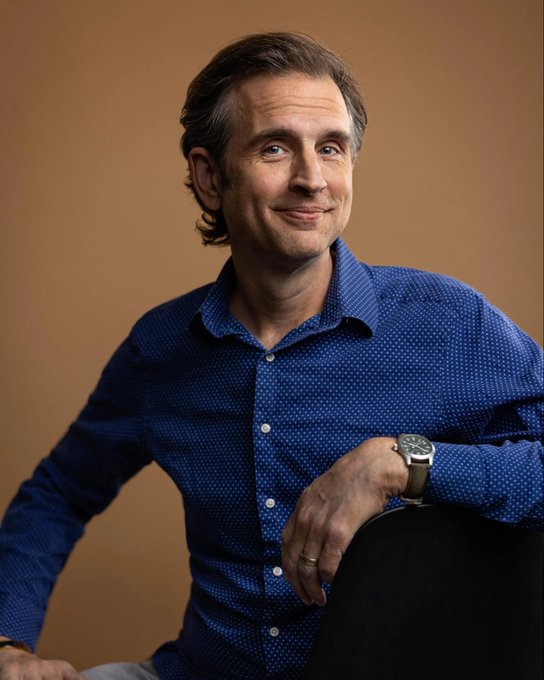 A man in a blue patterned button-up shirt sits smiling with arms crossed against a warm brown backdrop, wearing a silver wristwatch. The image appears to illustrate John, mentioned in the post as connected to the 2013 Zerocash research paper that laid the groundwork for ZCash ($ZEC).