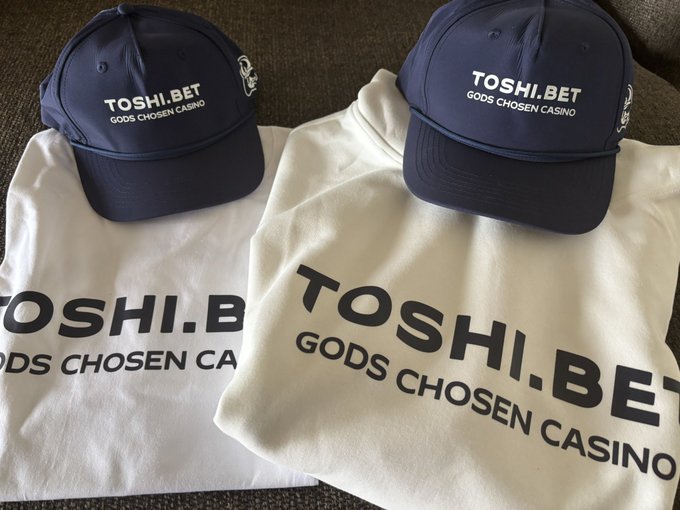 Two navy blue baseball caps and white hoodies branded with 'TOSHI.BET' and 'GODS CHOSEN CASINO' in black text, laid out on a surface.