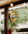 An oval illuminated sign for Shiner Beer hangs from a wooden beam of a covered patio, with trees and hanging lights visible in the background.