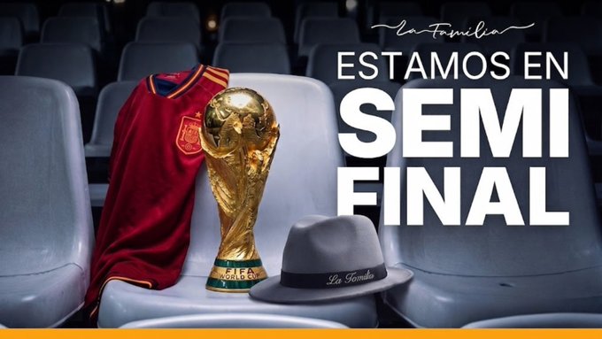 A Spain national football team jersey is draped over a stadium seat next to the FIFA World Cup trophy and a gray "La Familia" fedora, with bold text announcing "ESTAMOS EN SEMI FINAL" (We're in the Semifinal), celebrating Spain's advancement and promoting a July 14, 2026 community gathering in Madrid with Chiliz and the Solana ecosystem.